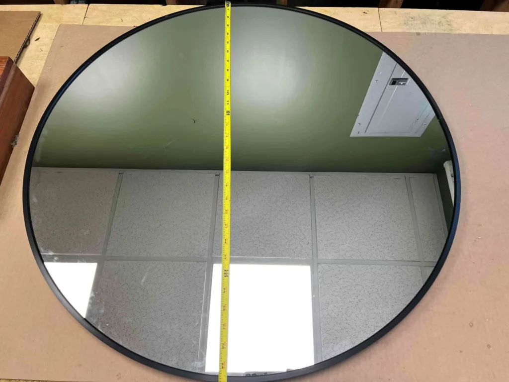 A round mirror with a black metal edge with a tape measure going from the upper side to the lower side of the mirror.