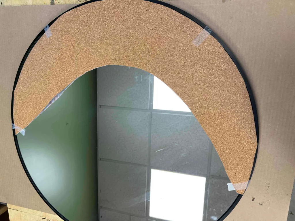 A round mirror with a black metal edge and a dark tan color crescent shape panel on one of the sides.