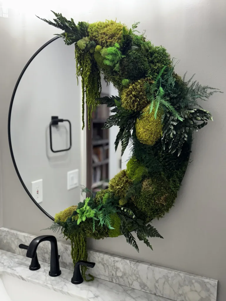 A round bathroom mirror with a black edge and on the right hand side there is a combination of different shades of green moss and plants.